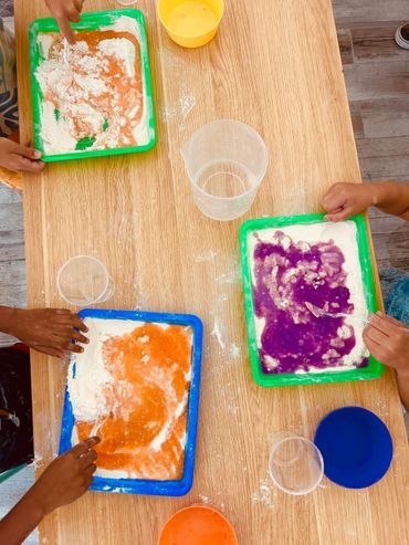 Children mixing colorful liquids with powder in trays on a wooden table.