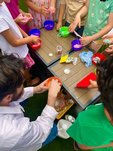 Children and an adult making colorful slime at a table outdoors.
