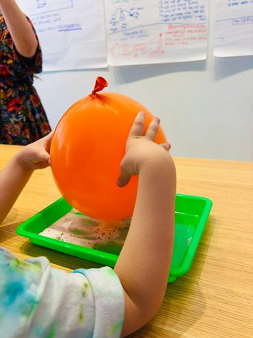 A child holds an orange balloon over a green tray with sand on a wooden table.