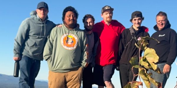 Group of six men posing outdoors on a sunny day with clear sky.