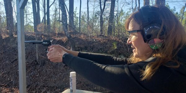 Woman wearing ear protection firing a handgun outdoors.