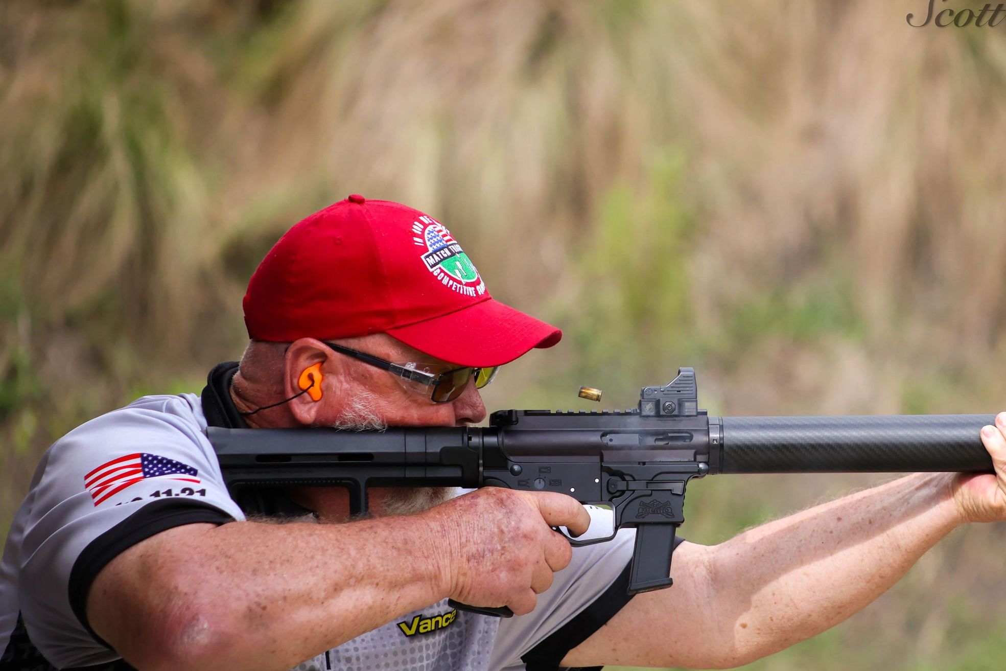 Man in red cap firing a rifle with ejected bullet in mid-air.