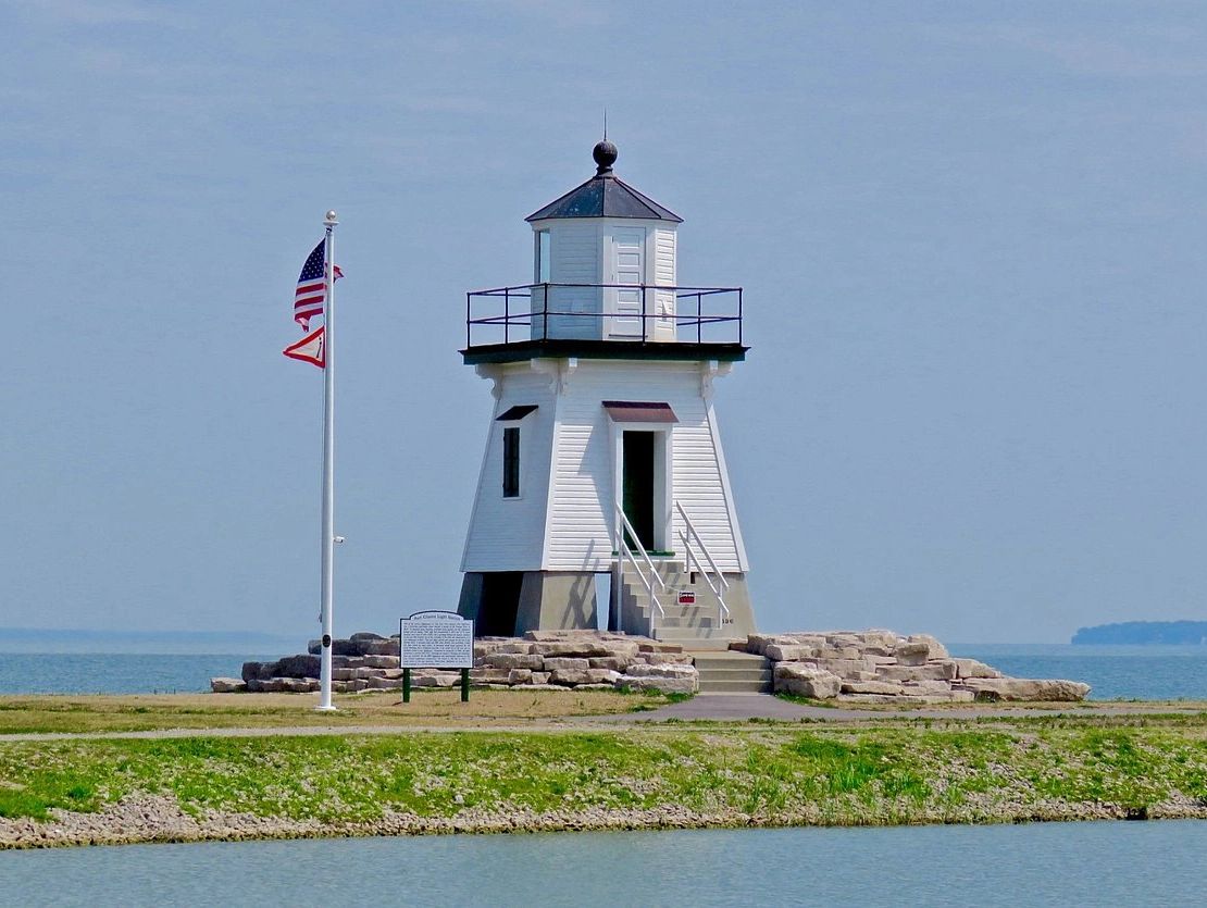 White lighthouse on a rocky shore with American flags flying beside it.