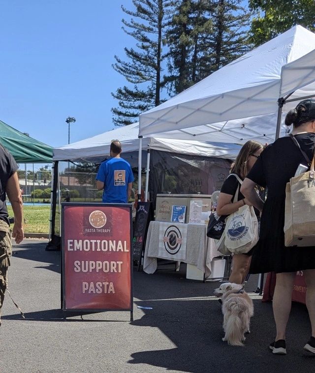 Outdoor market booth offering 'Emotional Support Pasta' with people and a dog nearby.