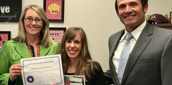 Three smiling adults holding a certificate and a book indoors.