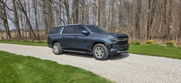 A dark gray Chevrolet Suburban parked on a gravel driveway by a woodland area.