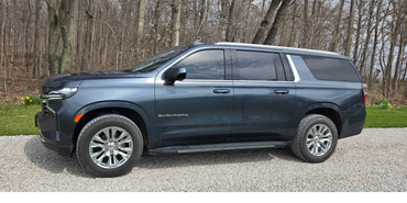 Dark blue Chevrolet Suburban parked on gravel with forest background.