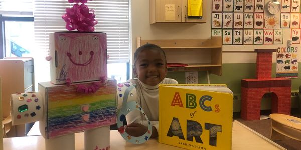 Child smiling with a colorful robot and art book in a classroom.