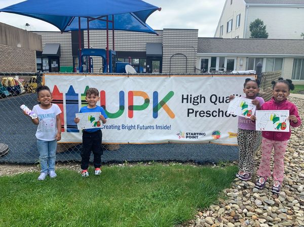 Four children smiling in front of a preschool banner holding artwork.