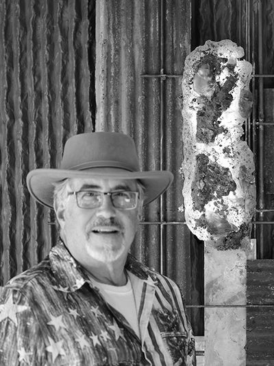 Man in patriotic shirt and hat stands before modern metal and glass sculpture.