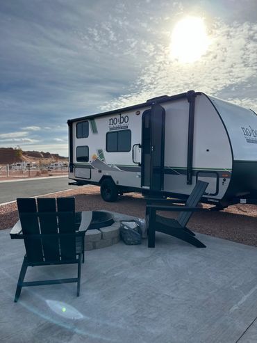 A camper trailer with two chairs and a fire pit under a bright sky.