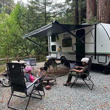 Family camping with kids and a dog near a camper trailer in the forest.