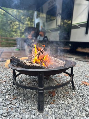 A fire pit with burning logs on gravel near an RV and blurred people.