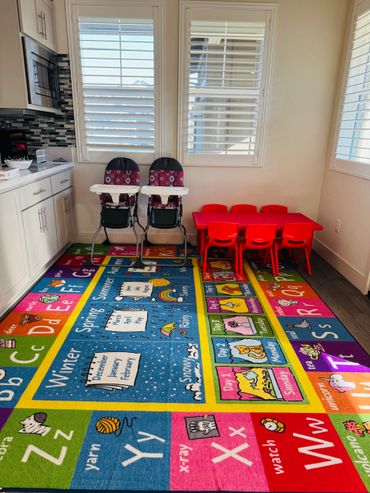 Colorful kids' play area with alphabet rug, red table, chairs, and two high chairs by windows.