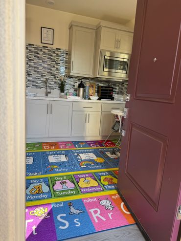 A colorful alphabet and days of the week rug in a small kitchen area with white cabinets.