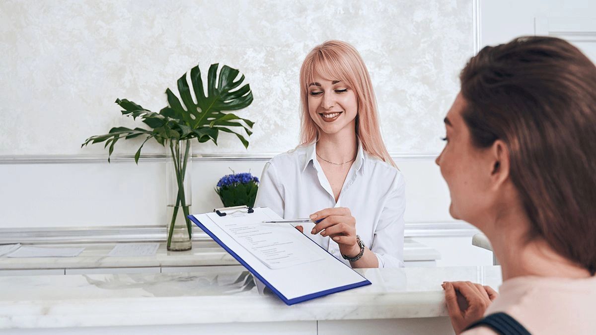 Receptionist smiling while showing a clipboard to a visitor.