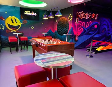Indoor mini golf and foosball game area with vibrant neon lights and cartoon murals.