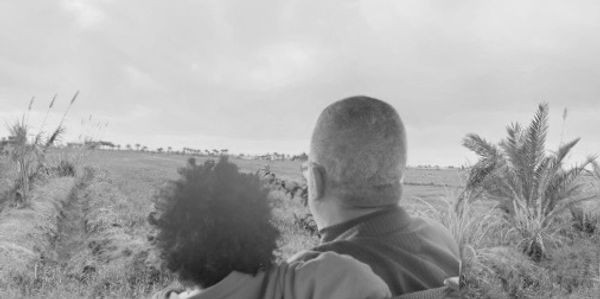 A couple embraces while looking at a vast field under a cloudy sky.