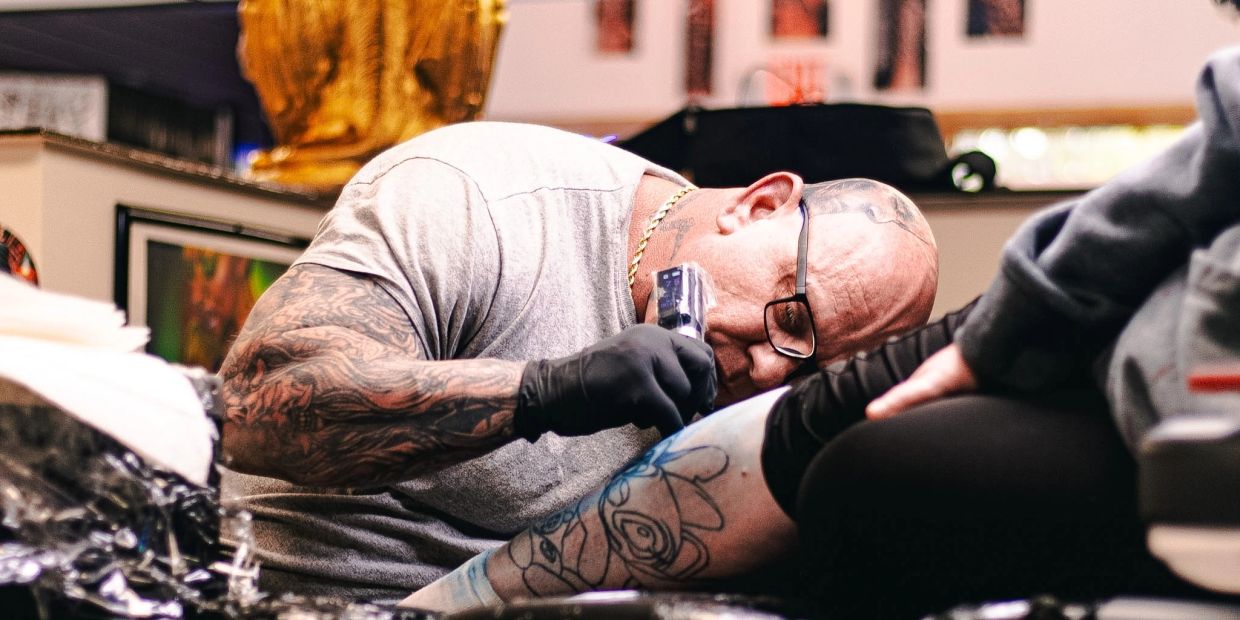 A tattoo artist carefully inks a blue cartoon design on a client's leg.