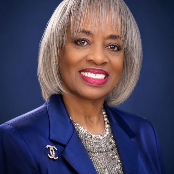 Elegant woman with silver hair smiles, wearing a blue blazer and statement necklace.