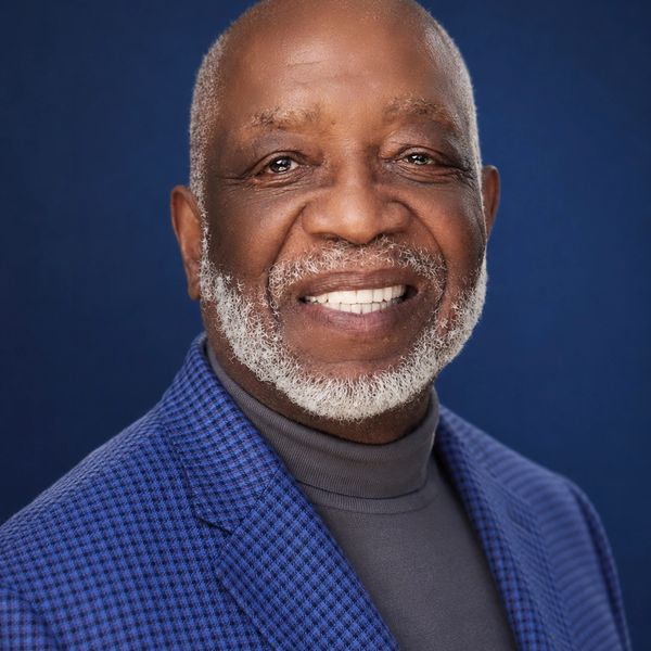 Smiling elderly man in a blue checkered blazer and gray turtleneck against a blue background.
