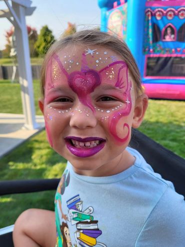 A child with vibrant unicorn face paint and purple lipstick smiles outdoors.