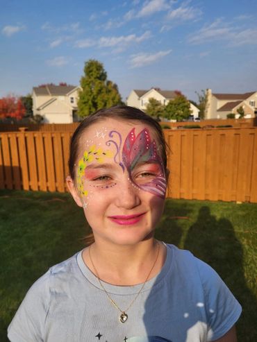 A girl with colorful butterfly face paint smiling outdoors on a sunny day.