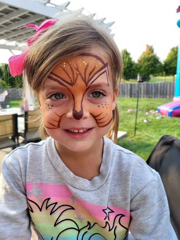 A young girl with a colorful cat face painting and a pink bow in her hair.
