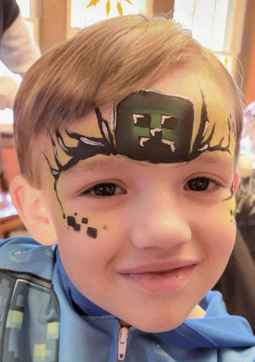Smiling child with Minecraft Creeper face paint and blue hoodie.