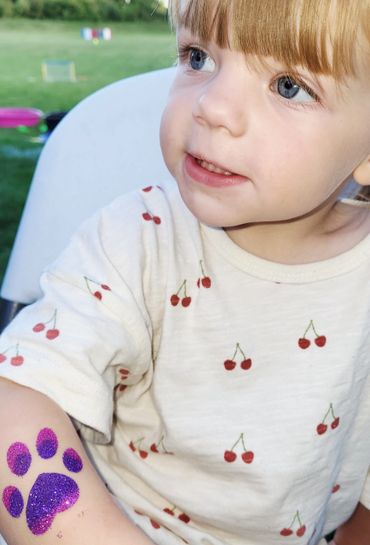 A child with blue eyes wearing a cherry-patterned shirt and a glittery purple paw print on their arm.