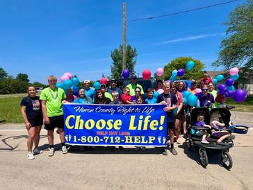 Group holding a 'Choose Life!' banner with colorful balloons on a sunny day.