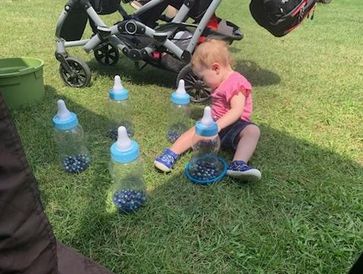 A toddler playing with blue marbles and baby bottles on grass near a stroller.