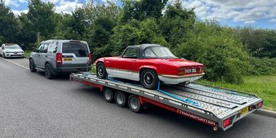 Red vintage sports car on a trailer hitched to a grey SUV on a country road.