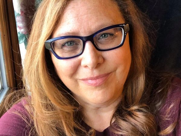 Smiling woman with glasses and long wavy hair near a window.
