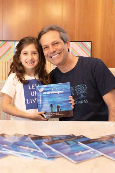 A smiling man and young girl proudly display a book together indoors.