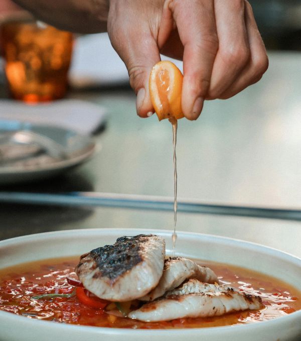 Hand squeezing citrus over grilled fish in tomato sauce.