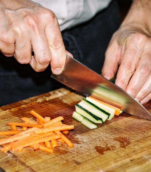 Hands slicing zucchini and carrots into thin sticks on a wooden cutting board.