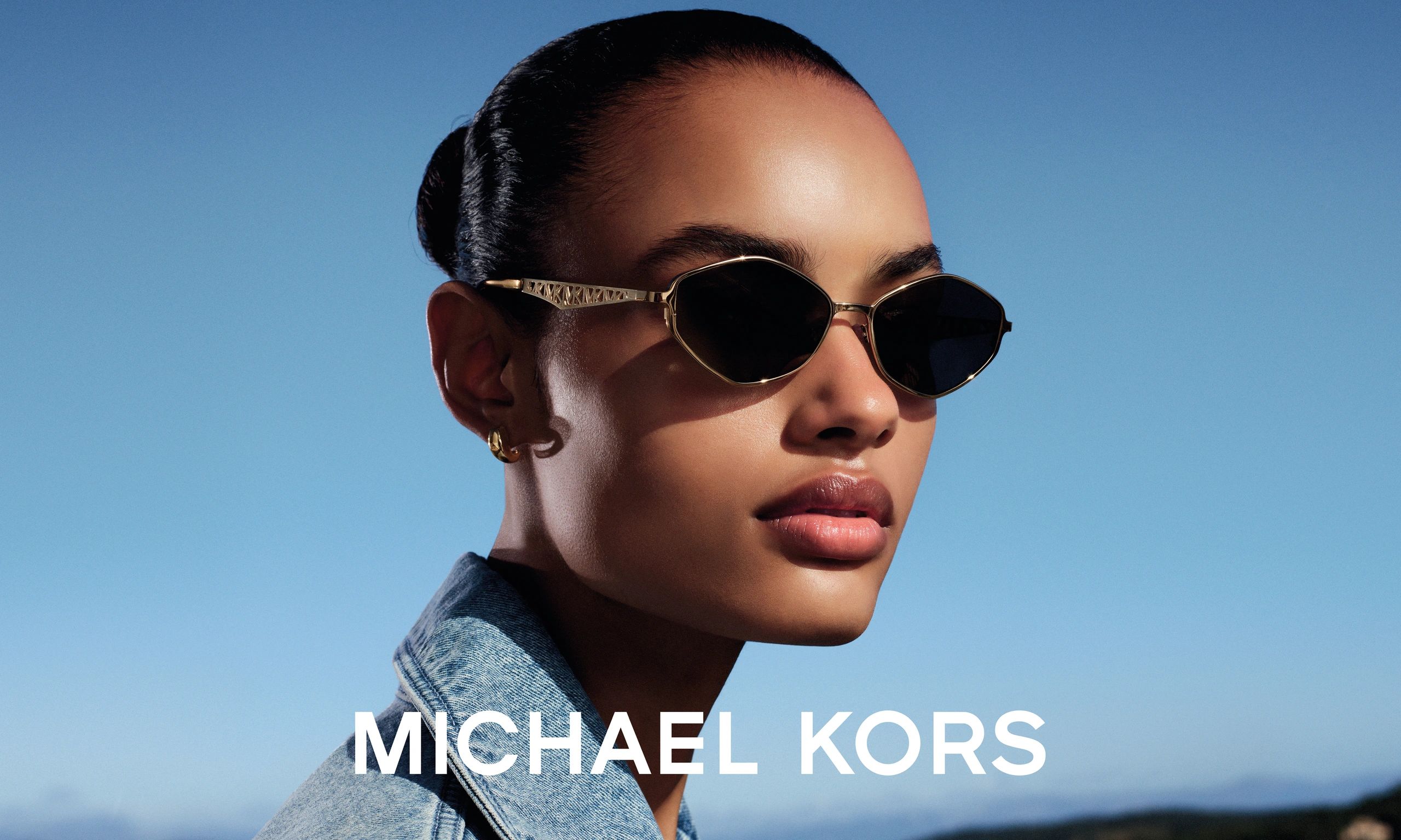 Stylish woman wearing Michael Kors sunglasses and a denim jacket against a clear blue sky.