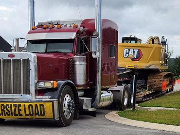 Red semi-truck carrying a large CAT excavator on a trailer with an oversize load sign.