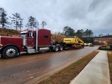 A red truck hauling a yellow Komatsu excavator on a trailer in a construction area.