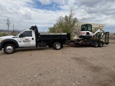 A white truck towing a trailer with a mini excavator on a dirt road.