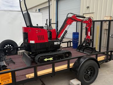 Compact red mini excavator on a flatbed trailer outside a warehouse.
