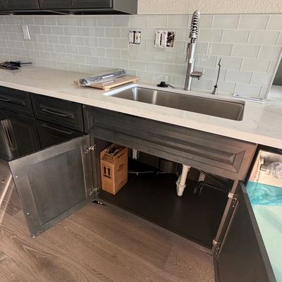 Open kitchen cabinet below sink with visible plumbing and a box inside.