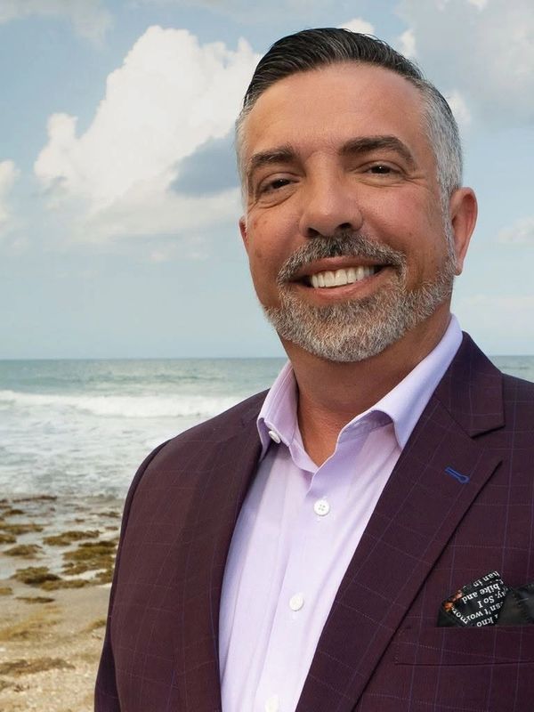 Smiling man in a suit standing by the ocean shore under a cloudy sky.