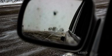 driver side mirror on a Jeep, with snow blowing across the road in the foreground and in the mirror