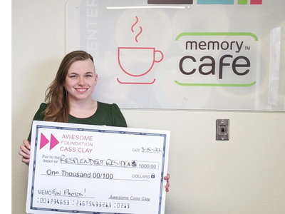 A smiling woman stands next to a memory cafe sign, holding a thousand dollar check.