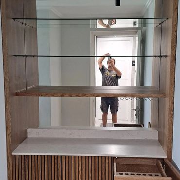 Man taking a selfie reflection in a wooden cabinet with glass shelves.