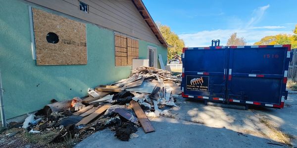 A pile of construction debris next to a house and a large blue dumpster.