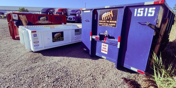 Three colorful dumpsters with company logos on a gravel lot under clear blue sky.