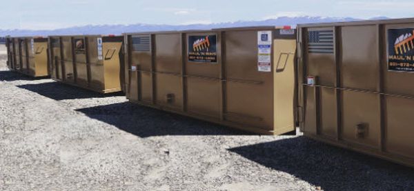 A row of brown industrial dumpsters lined up outdoors on gravel.